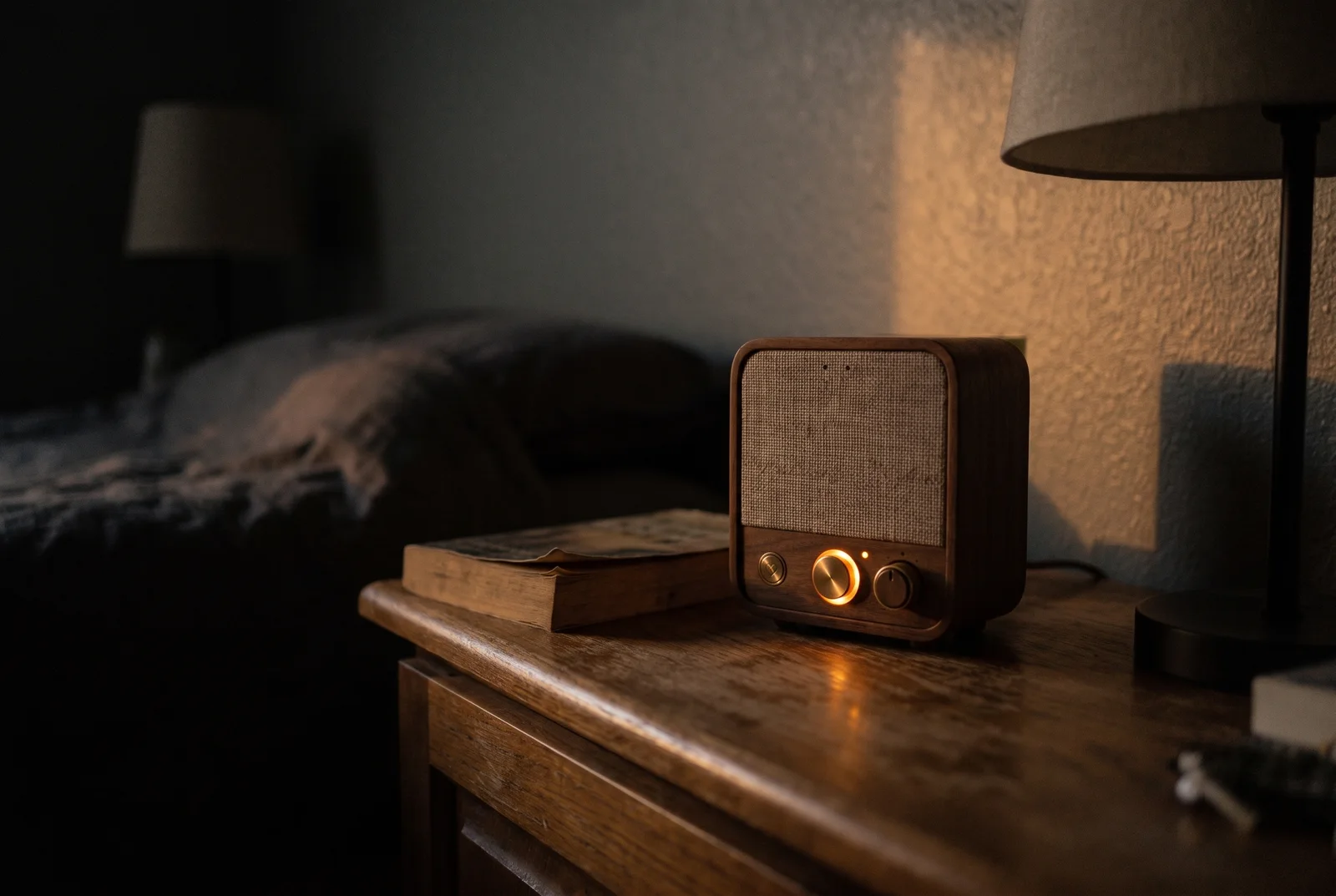 Roomie on a bedside table in a dark room at dusk, its amber ring the only light, throwing a warm glow up the wall beside a made bed and a stack of books.